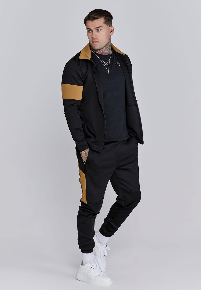 Black athletic tracksuit with gold accents, featuring a zip-up jacket and tapered pants. Worn with white sneakers, showcasing a fitted design.