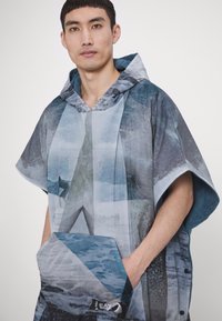 Hætteponcho med et blå-gråt fotografisk print, frontlomme og trykknaplukninger, lavet af let, tekstureret stof.
