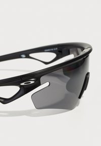 Sport Sonnenbrille mit schwarzem Rahmen, grauen Gläsern und offenem Seitendesign. Zu den Merkmalen gehören eine schlanke Form und markante Aussparungen an den Bügeln.