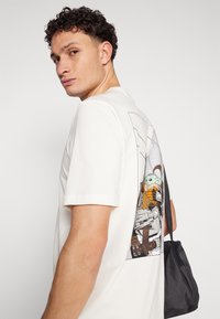 Βαμβακερή μπλούζα με μανίκια σε off-white χρώμα, με γραφικό τύπωμα στην πλάτη ενός χαρακτήρα που κρατάει ένα σνακ, συνδυασμένη με μια μικρή μαύρη τσάντα.