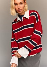 Rdeča polo majica z dolgimi rokavi in belimi ter mornarsko modrimi črtami, pod katero je bela srajca s ovratnikom. Tkanina se zdi mehka, elegantna in sproščena.