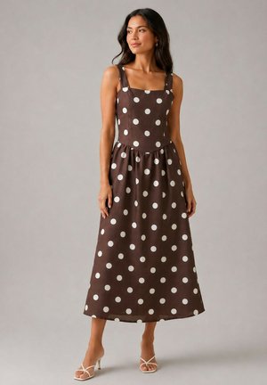 Femme portant une robe midi marron sans manches à pois blancs et encolure carrée, associée à des sandales blanches à talons, debout sur un fond gris.