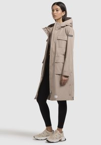 Beige vízálló parka kapucnival, elöl patentos zsebekkel, cipzáras záródással és állítható oldalsó szegélyekkel. Fekete leggings és tornacipő fölé viselve.