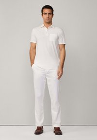 Bílý polo tričko s kapsou na hrudi, kombinované s bílými kalhotami a hnědými mokasíny. Oblečení má krátké rukávy a klasický límec.