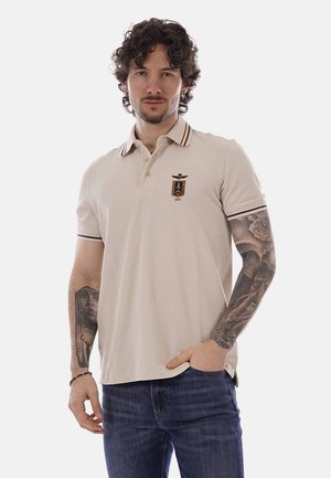 Uomo con capelli ricci e tatuaggi che indossa una polo beige con stemma e jeans blu, in piedi con una mano in tasca su sfondo bianco.