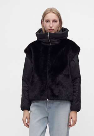 Femme aux cheveux blonds courts portant une veste sans manches en fausse fourrure noire par-dessus une doudoune noire et un jean bleu clair, debout devant un fond uni.