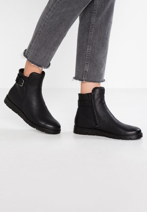 Person iført sorte ankelstøvler med sidelukning og spænde, kombineret med grå jeans med frynset kant på en hvid gulv.