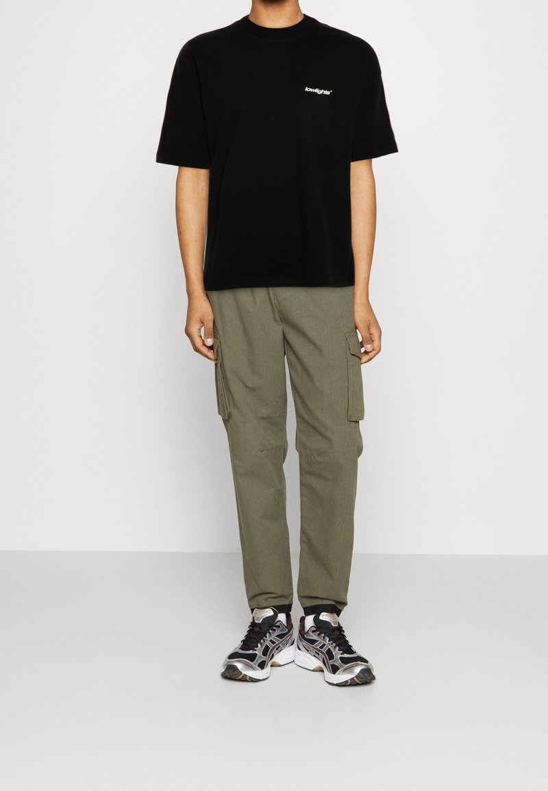 Czarna bawełniana koszulka z logo, oliwkowe spodnie cargo z bocznymi kieszeniami oraz czarne i srebrne buty sportowe. Prosty, casualowy zestaw.