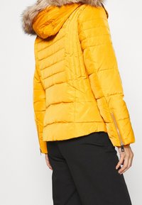 Giacca a vento gialla con cappuccio bordato di pelliccia, texture trapuntata, dettagli con zip laterali e silhouette aderente, indossata sopra pantaloni neri.