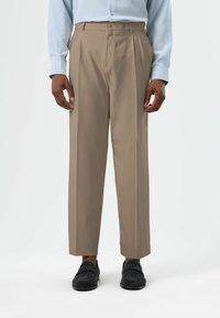 Beige pleated trousers with a relaxed fit, made from a smooth fabric, featuring a waistband and side pockets, paired with black shoes.