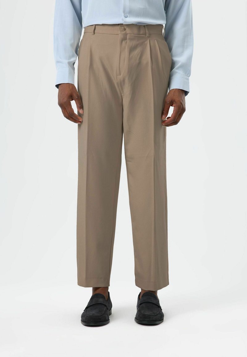Beige pleated trousers with a relaxed fit, made from a smooth fabric, featuring a waistband and side pockets, paired with black shoes.