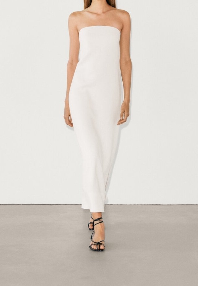 Femme portant une robe blanche longue sans bretelles et des sandales noires à talons hauts avec des lanières, debout contre un mur blanc uni.