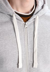 Felpa con zip grigia chiara e coulisse bianche. Caratterizzata da una texture liscia e una cerniera frontale. Indossata sopra una maglietta a strati marrone e nera.
