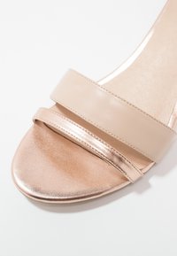 Sandale en cuir beige avec une lanière dorée rose brillante et un bout lisse et arrondi. Présente un design épuré et minimaliste avec une semelle plate.