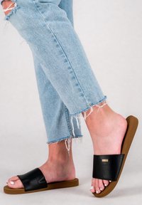 Schwarze, strukturierte Slidesandalen mit einer flachen braunen Sohle, getragen mit hellblauen, ausgefransten Jeans, wobei die Zehen sichtbar sind und minimalistische Designdetails hervorgehoben werden.