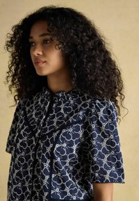 Tumedatest sinine pluus, millel on keerukas valge lilleline tikand, lühikesed puhv-sleeved varrukad ja sakilise kaelusega pitsi ääris.