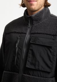 Veste en polaire noire avec un haut en tissu lisse et un bas texturé. Comprend des zips, une poche avant et un patch logo. Conception à col haut.