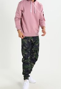 Sudadera rosa claro con cordones blancos, combinada con pantalones de camuflaje verde oscuro con puños. El modelo lleva zapatillas blancas.