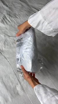 Borsa bianca contrassegnata "NEXT", realizzata in plastica riciclata, tenuta in mano. Include un'etichetta del prodotto e istruzioni per il riciclaggio. Lo sfondo è in tessuto testurizzato.