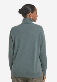 Kvinna med mörkt hår i en knut bär guld hoops, en blå polotröja och mönstrade byxor, sedd bakifrån mot en vit bakgrund.