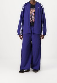 Lila träningsoverall med vita ränder längs ärmarna och sidorna, med en grafisk svart t-shirt under, ihop med svarta flip-flops.