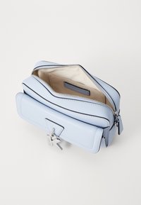 Světle modrá kožená taška se stříbrnými detaily, zipovým uzávěrem a béžovým podšívkou. Vyznačuje se plochým designem a přívěskem na klíče.