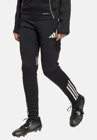 Schwarze Sporthosen mit weißen Seitenstreifen, AeroReady-Logo und einem reflektierenden Patch, kombiniert mit schwarzen Fußballschuhen.