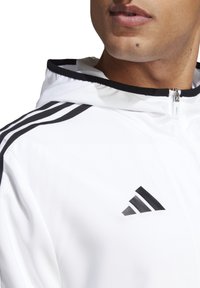 Chaqueta blanca con cremallera y acentos negros, que presenta tres franjas distintas en los hombros y un logotipo bordado en el pecho.
