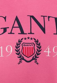 Růžové bavlněné tričko s logem "GANT" v námořnické modři, s designem červeného a bílého štítu a vyšívanými vavřínovými akcenty.