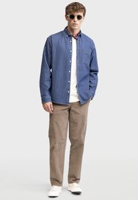Blue and white striped button-up shirt, white t-shirt underneath, beige pants, and white sneakers, accessorized with round black sunglasses.