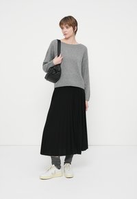 Kvinna som bär en grå tröja, svart plisserad kjol, grå benvärmare, vita sneakers och bär en svart axelväska.