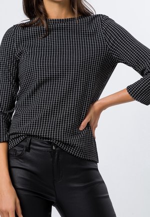 Frau trägt ein schwarz-weiß kariertes Langarmoberteil und schwarze Hose, mit einer Hand auf der Hüfte vor einem einfarbigen Hintergrund.