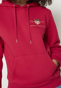 Czerwona bawełniana bluza z kapturem z przednią kieszenią, regulowanymi troczkami i haftowanym logo na piersi przedstawiającym tarczę i tekst.