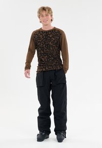 Braunes Langarmtop mit schwarzem Grafikdruck; kombiniert mit lockeren schwarzen Hosen und schwarzen Skistiefeln. Schlichtes Design, keine bemerkenswerten Akzente.