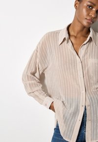 Camisa beige ligera de botones con sutiles rayas verticales, mangas largas y un bolsillo en el pecho, hecha de una tela suave y translúcida.