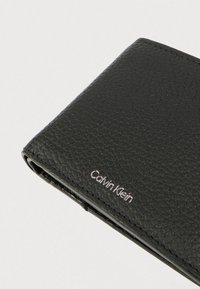 Μαύρο πορτοφόλι από υφή δέρματος με ορατή ραφή και ασημένιο ανάγλυφο λογότυπο "Calvin Klein" στην κάτω δεξιά γωνία.