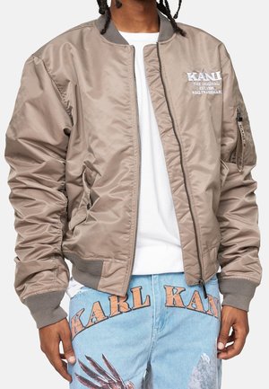 Person trägt eine beige Bomberjacke mit dem "KANI"-Logo auf der Brust und hellblaue Jeans mit dem Schriftzug "KARL KANI" und grafischem Druck.