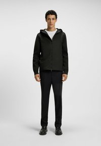 Svart zip-up jacka med huva, långa ärmar och två framfickor, kombinerad med svarta byxor och svarta skor. Slät textur överallt.