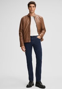 Homme portant une veste en cuir marron sur une chemise blanche, un jean bleu foncé et des bottes noires, se tenant avec une main dans la poche.
