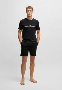 Schwarzes T-Shirt mit Rundhalsausschnitt und einem hellen Akzentband, auf dem das Wort "BOSS" steht. Kombiniert mit schwarzen Shorts, auf einem neutralen Hintergrund präsentiert.