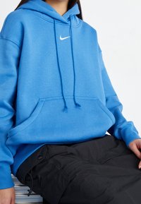 Blå hoodie med dragsko och känguruficka. Har en liten vit Nike-swoosh-logga på bröstet. Mjuk tyg och avslappnad passform.