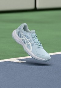 Lyseblå sportssko med tekstureret finish, snørelukning og hvide detaljer, vist i luften over en baneoverflade.