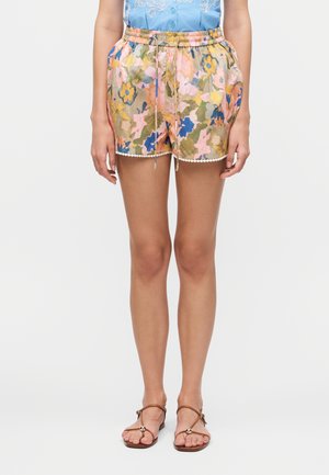 Nő virágmintás rövidnadrágot visel gumis derékrésszel és zsinórral, kék blúzzal és barna szandállal párosítva.