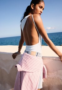 Un body sans dos bleu clair associé à un short en denim clair. Une chemise rayée rose et blanche est nouée à la taille. L'océan en arrière-plan.