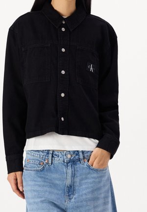 Kobieta ubrana w czarną krótką kurtkę na guziki na białą koszulę oraz jasnoniebieskie jeansy, z ręką w kieszeni, stojąca na jednolitym tle.