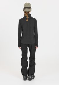 Schwarzes Langarm-Unterhemd mit hohem Kragen, kombiniert mit lockeren schwarzen Hosen. Eine Strickmütze mit einem hellen und dunklen Muster rundet das Outfit ab.