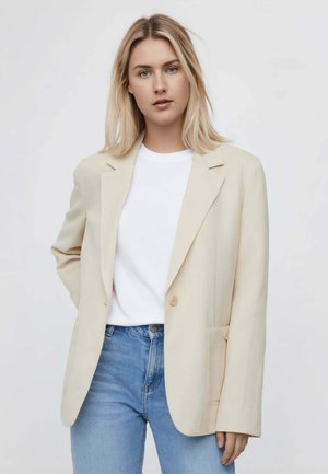 Giovane donna con capelli biondi che indossa un blazer beige sopra una camicia bianca e jeans blu, in piedi davanti a uno sfondo chiaro e uniforme.