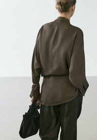 Frau mit Hochsteckfrisur, die eine locker sitzende braune Bluse und eine Hose trägt, eine kleine schwarze Handtasche hält und vor einem schlichten weißen Hintergrund steht.