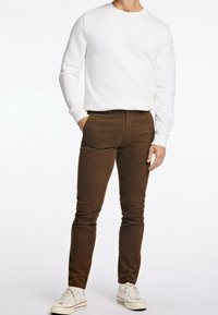 Homme portant un sweat-shirt blanc à manches longues et un pantalon en velours côtelé marron avec des baskets blanches, debout avec une main dans la poche sur fond blanc.