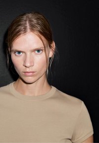 Béžové tričko s kulatým výstřihem vyrobené z bavlny, nošené osobou se rovnými vlasy na černém pozadí. Minimalistický design, hladká textura.